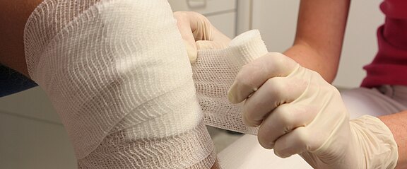 Nahaufnahme: Eine Person mit Einweghandschuhen legt einem verletzten Knie einen dicken Verband an. Weiße Mullbinden werden sorgfältig um das Gelenk gewickelt. Szene vermittelt medizinische Versorgung und Wundbehandlung.