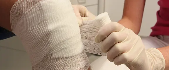Nahaufnahme: Eine Person mit Einweghandschuhen legt einem verletzten Knie einen dicken Verband an. Weiße Mullbinden werden sorgfältig um das Gelenk gewickelt. Szene vermittelt medizinische Versorgung und Wundbehandlung.