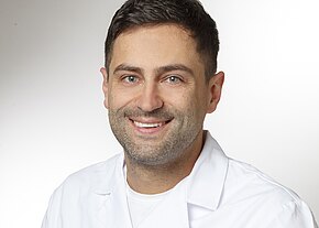 Dr. med. Christian Feichter Portrait eines lächelnden Mannes in einem weißen Kittel. Er hat kurze, dunkle Haare und trägt ein Unterhemd. Der Hintergrund ist hell und sanft und vermittelt eine freundliche Atmosphäre.