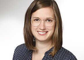 Porträt einer lächelnden Person mit runder Brille und schulterlangem braunem Haar. Die Person trägt ein dunkelblaues, weiß gepunktetes Hemd vor hellem, neutralem Hintergrund.