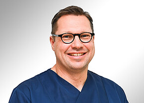 Dr. med. Rainer Getto Porträt eines lächelnden Mannes mit runder schwarzer Brille und kurz geschnittenen Haaren. Er trägt dunkelblaue medizinische Kleidung und blickt direkt in die Kamera vor hellem, neutralem Hintergrund.