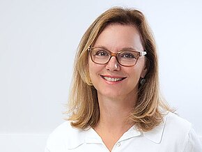 Freundlich lächelnde Frau mit Brille sitzt an einem Schreibtisch, Hände an Computermaus und Tastatur. Auf dem Tisch liegt ein Stethoskop, Hinweis auf ein medizinisches Umfeld.