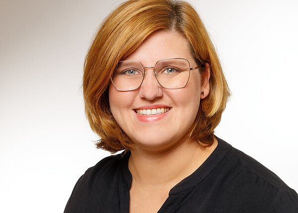 Porträt einer lächelnden Frau mit schulterlangem, rötlich-blondem Haar und großer, eckiger Brille. Sie trägt ein schwarzes Oberteil und schaut direkt in die Kamera vor hellem, neutralem Hintergrund.