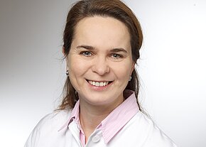 Dr. med. Claudia Anhut Porträt einer lächelnden Frau mit langen, braunen Haaren, die ein weißes Hemd mit rosa Kragen trägt. Der Hintergrund ist hell und neutral gehalten. Die Frau strahlt Freundlichkeit und Professionalität aus.