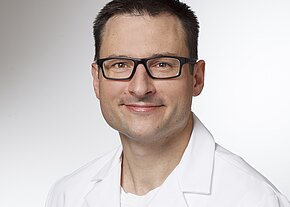Dr. med. Mark Schmolze Porträtfoto eines lächelnden Mannes mit schwarzer Brille und kurzem dunklem Haar, in einem weißen Kittel vor neutralem Hintergrund.