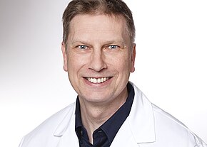 Porträt eines lächelnden Mannes mittleren Alters in weißem Kittel vor hellem Hintergrund; Kopf-und-Schulteraufnahme, direkter Blick in die Kamera, freundlicher, professioneller Eindruck.