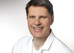 Dr. med. André Friedrich Porträtfoto eines mittelalten Mannes mit kurzen dunklen Haaren, der freundlich lächelt. Er trägt einen weißen Kittel vor neutral hellem Hintergrund.
