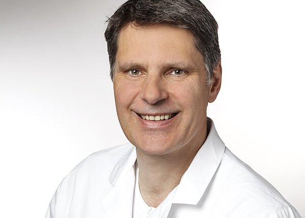 Porträtfoto eines mittelalten Mannes mit kurzen dunklen Haaren, der freundlich lächelt. Er trägt einen weißen Kittel vor neutral hellem Hintergrund.