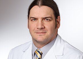 Dr. med. Lothar Veits Porträt eines Mannes mit zurückgebundenen dunklen Haaren. Er trägt einen weißen Kittel, graues Hemd und gestreifte Krawatte. Neutrale Mimik, Blick in die Kamera, heller Hintergrund. Wirkt wie ein medizinischer Fachmann.