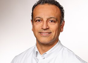 Ein lächelnder Mann trägt einen weißen Kittel. Sein Gesicht ist freundlich und er hat dunkle, leicht wellige Haare. Der Hintergrund ist schlicht und hell. Die Darstellung vermittelt einen professionellen Eindruck, der häufig mit medizinischen Berufen assoziiert wird.