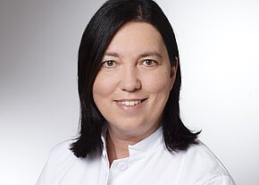 Ingrid Hegenbarth Porträt einer lächelnden Person mit schulterlangem, dunklem Haar im weißen Arztkittel vor neutralem Hintergrund.