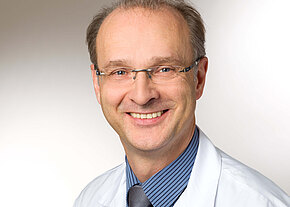 Prof. Dr. med. Jochen Willner Porträt eines freundlich lächelnden Mannes mittleren Alters mit Brille. Er trägt Hemd, Krawatte und einen weißen Kittel vor neutralem Hintergrund.