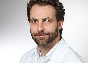 Gerson Schütze Porträtfoto eines bärtigen Mannes mit dunklem Haar in weißem Kittel, leichtes Lächeln, neutraler Hintergrund, Blick direkt in die Kamera.