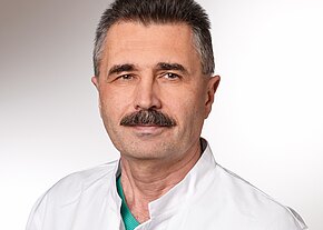 Detelin Dimov Ein männlicher Arzt mit Schnurrbart trägt einen weißen Kittel und ein grünes Oberteil. Er blickt freundlich in die Kamera. Der Hintergrund ist neutral und hell, was eine professionelle Atmosphäre vermittelt.