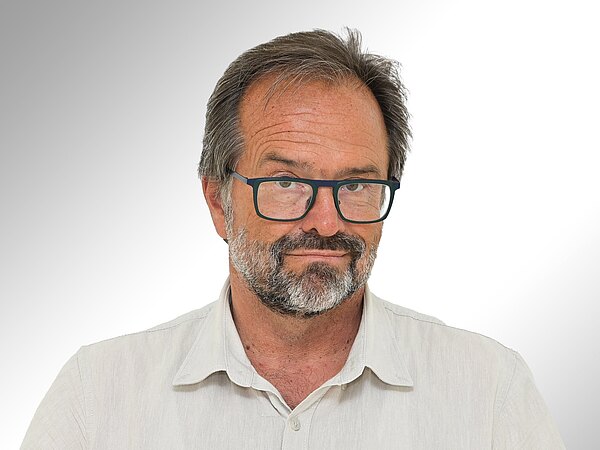 Ein Mann mit einem kurzen, grauen Bart und einer Brille blickt direkt in die Kamera. Er trägt ein helles, khakifarbenes Hemd. Der Hintergrund ist hell und verschwommen, was den Fokus auf sein Gesicht lenkt.