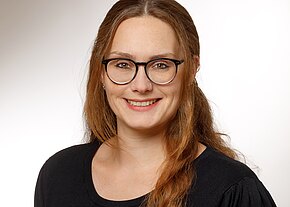 Madeleine Schmitt Porträtfoto: Eine lächelnde Frau mit runder schwarzer Brille und langen, leicht gewellten, brünetten Haaren. Sie trägt ein schwarzes Oberteil. Neutraler, heller Hintergrund.