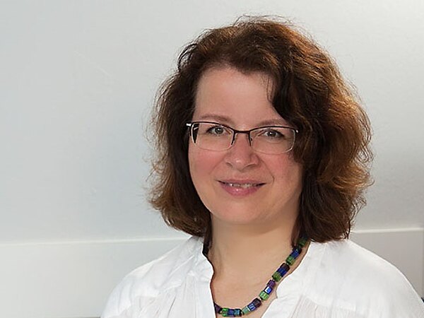 Freundlich lächelnde Frau mit Brille sitzt im Büro am Schreibtisch, trägt eine weiße Bluse, hält einen Stift und schaut in die Kamera.