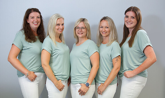 Fünf Frauen stehen in einer Reihe vor neutralem Hintergrund, tragen mintgrüne Oberteile und weiße Hosen und lächeln freundlich in die Kamera. Teamfoto.