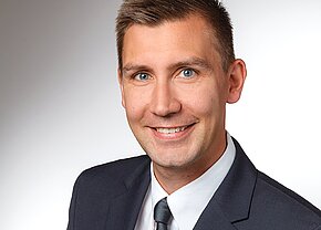 Hannes Diener MBA Ein lächelnder Mann trägt einen dunklen Anzug und eine hellere Krawatte. Der Hintergrund ist hell und einfarbig. Der Mann hat kurze, blonde Haare und blaue Augen.