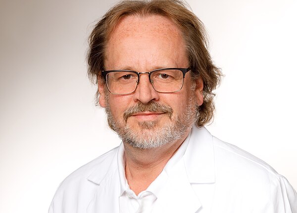 Frontales Porträt eines mittelalten Mannes mit Brille, kurzen welligen Haaren und grauem Bart. Er trägt einen weißen Kittel und Hemd und blickt ruhig in die Kamera vor hellem, neutralem Hintergrund.