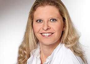 Porträtfoto: lächelnde erwachsene Frau mit langen, welligen, blonden Haaren. Sie blickt direkt in die Kamera. Neutraler, hellgrauer Studiohintergrund. Sie trägt weiße Oberbekleidung und kleine Ohrstecker.