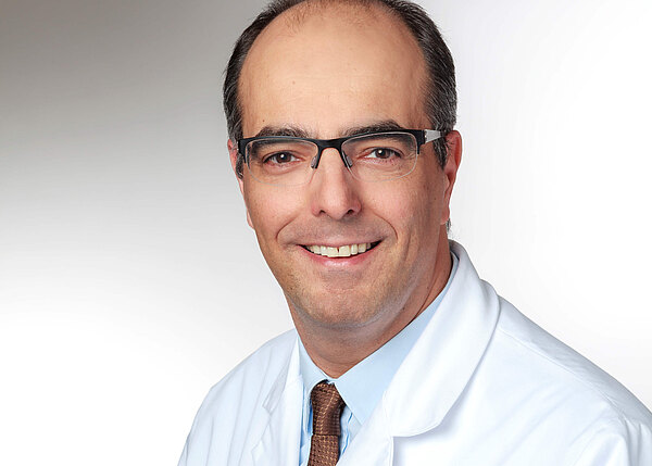 Porträt eines mittelalten Mannes mit Brille, der freundlich in die Kamera lächelt. Er trägt einen weißen Kittel, hellblaues Hemd und braune Krawatte. Neutraler, heller Hintergrund.