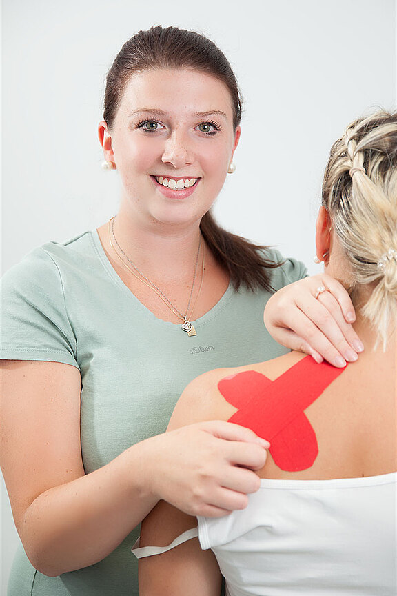 Therapeutin klebt rotes Kinesio-Tape auf die rechte Schulter und den oberen Rücken einer Patientin.