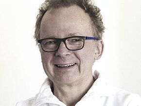 Porträt eines lächelnden Mannes mit Brille, weißes Oberteil, heller Hintergrund.